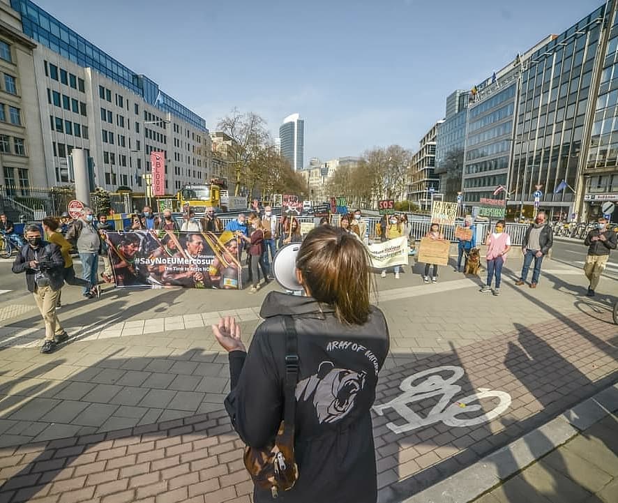 A person in an Army of Nature jacket holding a megaphone, facing a street demonstration behind barriers with banners reading Say No EU Mercosur