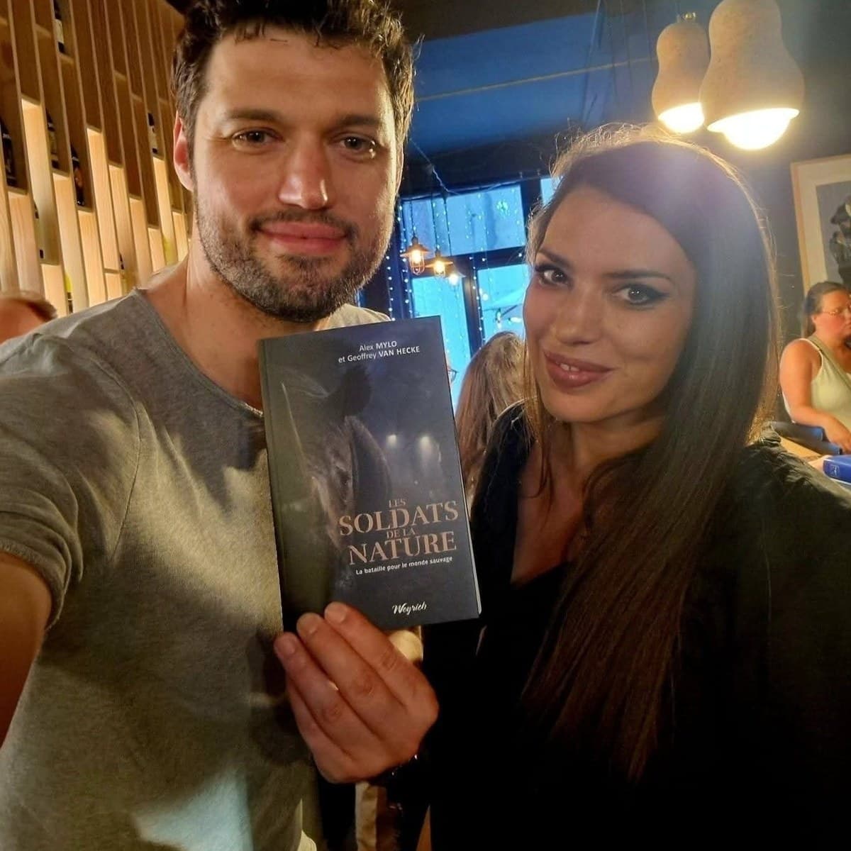 Two people at a book launch, one holding up a copy of Les Soldats de la Nature to the camera