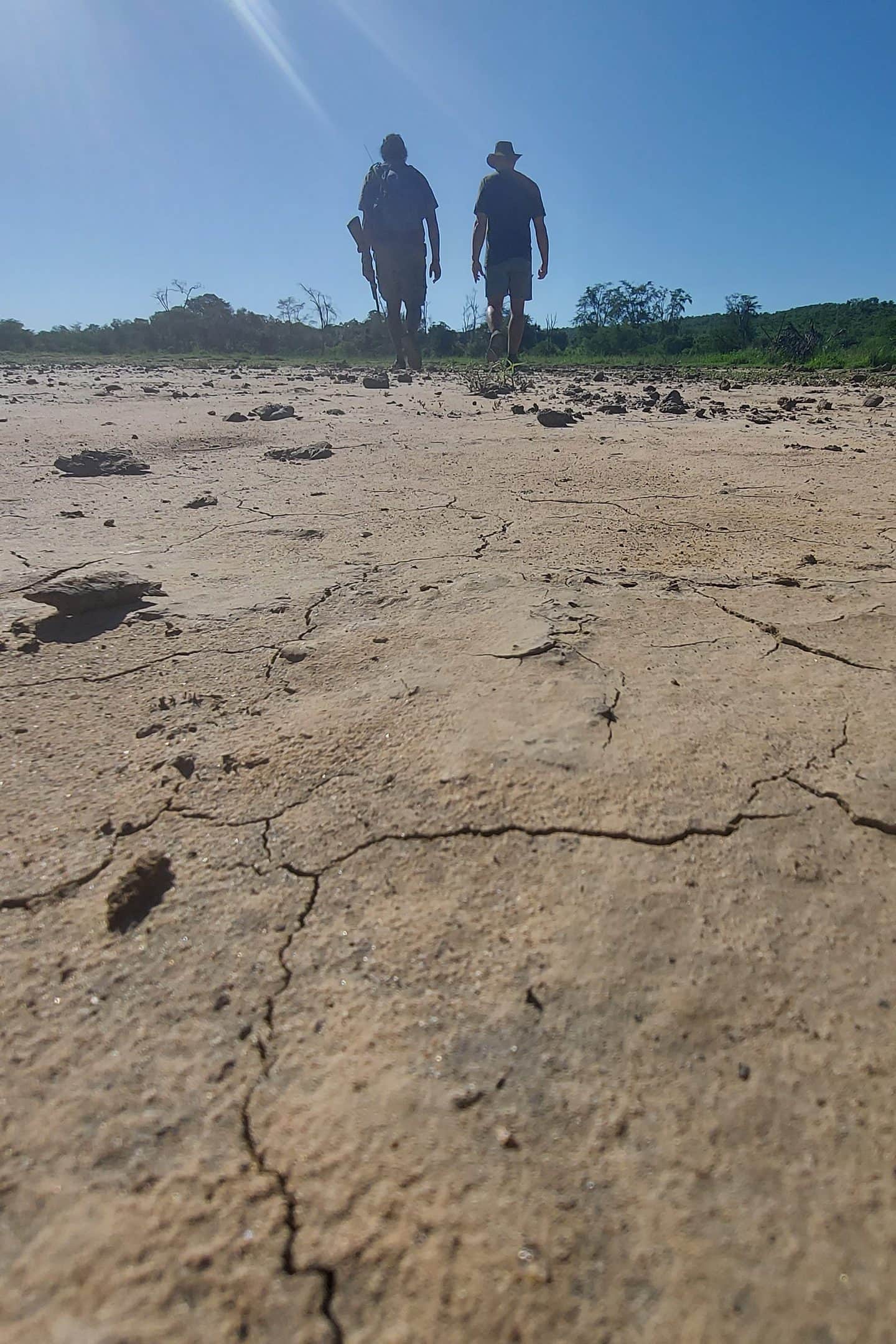 Two people walking up the middle of a dried-out riverbed, its clay floor cracked into plates