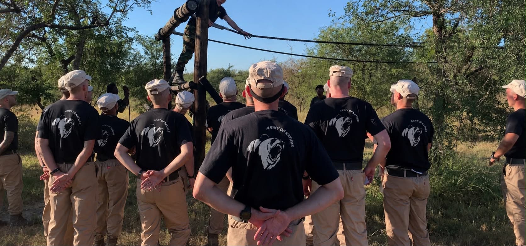 Trainees in Army of Nature T-shirts and field caps watching an instructor cross a rope traverse on a pole obstacle course, bushveld behind